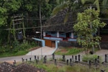 A traditional house with a sloping, tiled roof situated in a lush, green environment. The house features a veranda with a blue and white façade. There is a small concrete path leading to a closed gate. Surrounding the house are tall trees and foliage, and an electricity pole is visible on the left side.