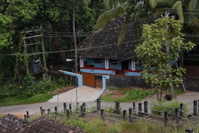 A traditional house with a sloping, tiled roof situated in a lush, green environment. The house features a veranda with a blue and white façade. There is a small concrete path leading to a closed gate. Surrounding the house are tall trees and foliage, and an electricity pole is visible on the left side.