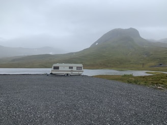 A solitary caravan is parked on a gravel area near a still water body, surrounded by green hills under an overcast sky. The landscape appears remote and tranquil, with a misty atmosphere emphasizing isolation.