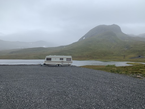 A solitary caravan is parked on a gravel area near a still water body, surrounded by green hills under an overcast sky. The landscape appears remote and tranquil, with a misty atmosphere emphasizing isolation.