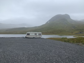 A solitary caravan is parked on a gravel area near a still water body, surrounded by green hills under an overcast sky. The landscape appears remote and tranquil, with a misty atmosphere emphasizing isolation.