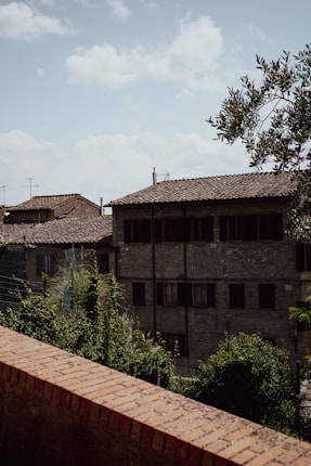 A rustic Italian village scene with old stone buildings featuring tiled roofs. A brick wall runs parallel to a paved street lined with greenery and vines. The sky is clear with few clouds, creating a serene atmosphere.