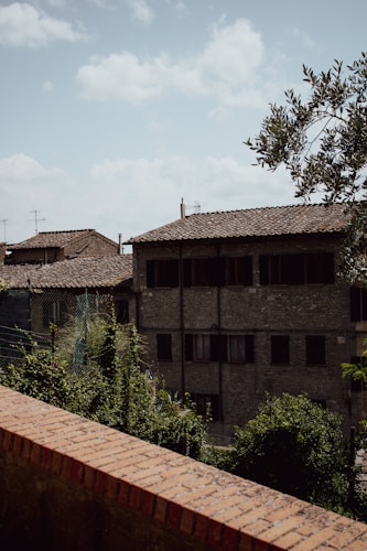 A rustic Italian village scene with old stone buildings featuring tiled roofs. A brick wall runs parallel to a paved street lined with greenery and vines. The sky is clear with few clouds, creating a serene atmosphere.