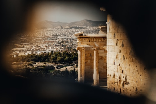 An ancient architectural structure with large stone columns and beige stone walls overlooks a sprawling cityscape. The city is densely packed with buildings and surrounded by rolling hills in the background. Tall trees can be seen in the foreground, adding a touch of greenery to the scene.