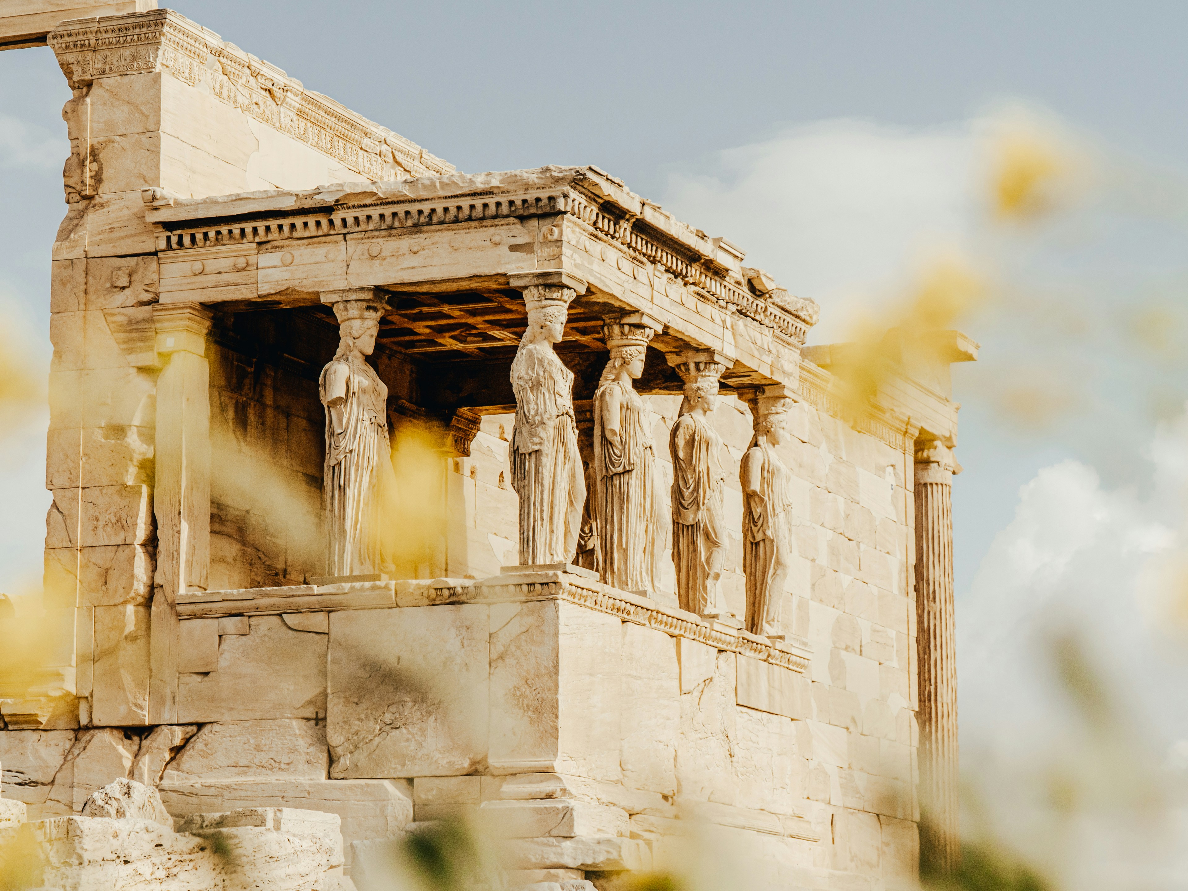 a group of statues on the side of a building, The image shows a detailed view of the Erechtheion, an ancient Greek temple on the Acropolis of Athens. The focus is on the Porch of the Caryatids, featuring six beautifully carved female statues (Caryatids) serving as architectural supports. The stone structure is bathed in warm, golden sunlight, highlighting the intricate details and weathered texture of the marble. The sky is clear with a few soft clouds, adding a serene backdrop to the historical monument. Yellow flowers in the foreground are slightly out of focus, adding a touch of natural beauty to the scene.