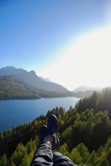 A pair of feet in hiking shoes is seen from a high vantage point overlooking a scenic landscape. The view includes a serene lake surrounded by dense forests, with distant mountains under a clear blue sky. The sunlight casts a bright glow over the horizon, creating a mixture of light and shadows across the landscape.