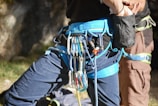 Two people are wearing rock climbing harnesses. Various climbing gear, including carabiners and quickdraws, are attached to one of the harnesses. The background features an outdoor setting with rock surfaces partially in the frame, and some grassy patches are visible.