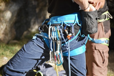 Two people are wearing rock climbing harnesses. Various climbing gear, including carabiners and quickdraws, are attached to one of the harnesses. The background features an outdoor setting with rock surfaces partially in the frame, and some grassy patches are visible.
