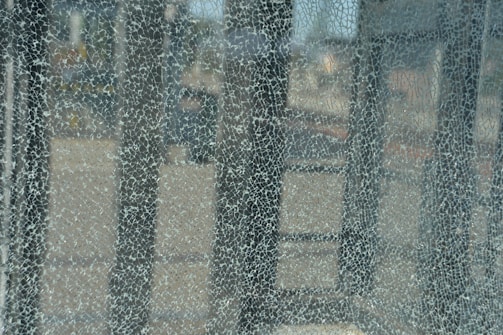 A close-up view of a shattered glass surface, revealing a network of intricate cracks and fragmented patterns. The background appears blurred, with indistinct shapes and colors visible through the broken glass.