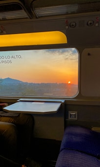 A train interior showing a window with a scenic view of a sunset over a city. There is a partially visible person sitting on a blue seat next to a small foldable table. The sky outside is a blend of orange and blue hues, and a hill or mountain is visible in the distance.
