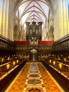 A grand, ornate cathedral interior with high vaulted ceilings featuring pointed arches. The central aisle is lined with wooden choir stalls, and there are elegant chandeliers emitting a warm glow. An impressive pipe organ is installed at the end of the aisle, adding to the majestic atmosphere.