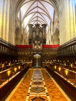 A grand, ornate cathedral interior with high vaulted ceilings featuring pointed arches. The central aisle is lined with wooden choir stalls, and there are elegant chandeliers emitting a warm glow. An impressive pipe organ is installed at the end of the aisle, adding to the majestic atmosphere.
