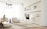 A minimalist living room with neutral tones featuring a woman sitting on a plush chair. The room includes modern decor, a wooden table with vases on built-in shelves, and a contemporary TV on a slim stand. Light flows through large, sheer drapes, creating a soft and serene atmosphere.