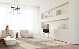 A minimalist living room with neutral tones featuring a woman sitting on a plush chair. The room includes modern decor, a wooden table with vases on built-in shelves, and a contemporary TV on a slim stand. Light flows through large, sheer drapes, creating a soft and serene atmosphere.