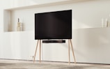 A modern TV on a minimalist wooden stand set against a light-colored wall. Decorative items like vases rest on shelves in the background, and there is a carpeted floor beneath.