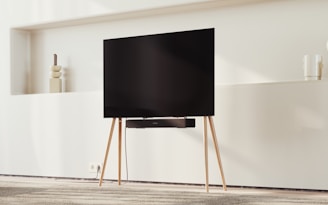 A modern TV on a minimalist wooden stand set against a light-colored wall. Decorative items like vases rest on shelves in the background, and there is a carpeted floor beneath.