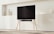 A modern TV on a minimalist wooden stand set against a light-colored wall. Decorative items like vases rest on shelves in the background, and there is a carpeted floor beneath.