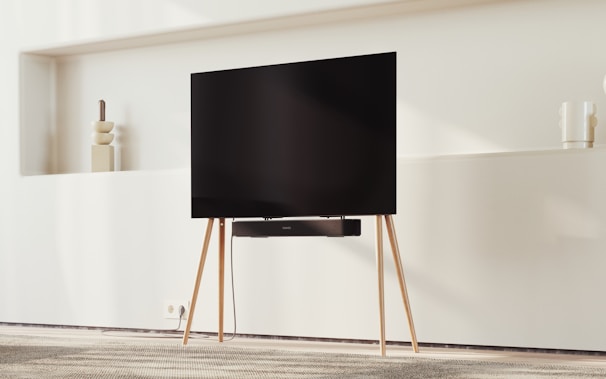 A modern TV on a minimalist wooden stand set against a light-colored wall. Decorative items like vases rest on shelves in the background, and there is a carpeted floor beneath.