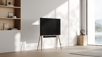 A minimalist, modern living space featuring a thin flat-screen TV mounted on wooden legs. Next to the TV, there's a small wooden table with a textured sphere on top. To the left is a built-in wooden shelf holding books and decorative items. Sunlight pours in from a large window overlooking a scenic view with mountains and a lake. The interior is bright with neutral tones, emphasizing simplicity and elegance.