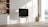 A minimalist, modern living space featuring a thin flat-screen TV mounted on wooden legs. Next to the TV, there's a small wooden table with a textured sphere on top. To the left is a built-in wooden shelf holding books and decorative items. Sunlight pours in from a large window overlooking a scenic view with mountains and a lake. The interior is bright with neutral tones, emphasizing simplicity and elegance.