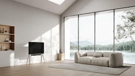 A minimalist living room with large floor-to-ceiling windows offering a scenic view of a lake and mountains. The room contains a comfortable cream-colored sofa set on a textured rug. A sleek television stands on a wooden tripod, and built-in shelves hold neatly arranged books and decorative items. Natural light floods the room, enhancing the serene atmosphere.
