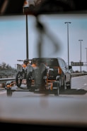 A car is towing a trailer with two off-road motorcycles on a highway. The scene is captured from a low angle, possibly from another vehicle, showing clear skies and street lamps along the road.