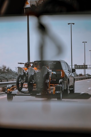 A car is towing a trailer with two off-road motorcycles on a highway. The scene is captured from a low angle, possibly from another vehicle, showing clear skies and street lamps along the road.