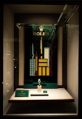 A store display case featuring a Rolex watch on a stand. Behind the watch, there is a vibrant, geometric cityscape design with tall, colorful buildings and the Rolex logo at the top. The display is well-lit, highlighting the watch and the decorative backdrop.
