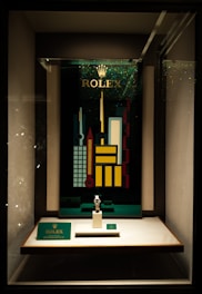 A store display case featuring a Rolex watch on a stand. Behind the watch, there is a vibrant, geometric cityscape design with tall, colorful buildings and the Rolex logo at the top. The display is well-lit, highlighting the watch and the decorative backdrop.