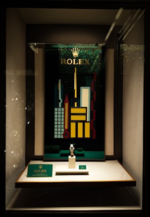 A store display case featuring a Rolex watch on a stand. Behind the watch, there is a vibrant, geometric cityscape design with tall, colorful buildings and the Rolex logo at the top. The display is well-lit, highlighting the watch and the decorative backdrop.