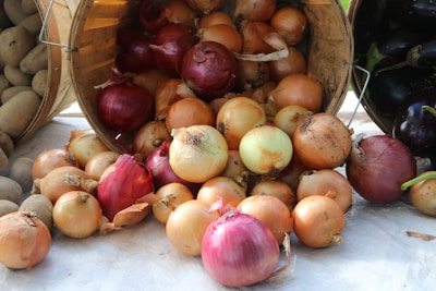 A collection of onions is scattered on a surface, coming out of a wooden basket. The onions vary in color, including yellow, white, and red. Nearby, there are also potatoes in another basket.