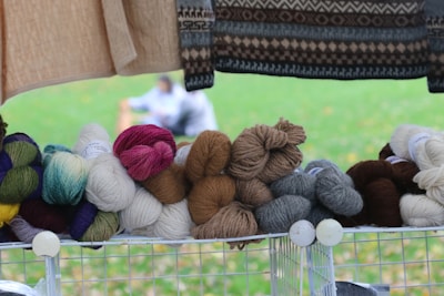 A variety of colorful yarn balls stacked neatly on a shelf. In the background, a blurred outdoor scene is visible, possibly of people sitting on grass. A knitted sweater with a brown and black pattern hangs above the yarn.