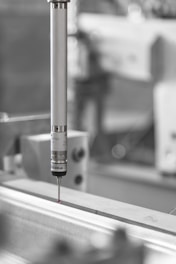 A precision measuring probe is positioned above a metallic surface, likely part of an industrial machine or CNC equipment. The focus is primarily on the metallic probe with a small red tip, highlighting its use in detailed measurements. The background is blurred, emphasizing an industrial setting.