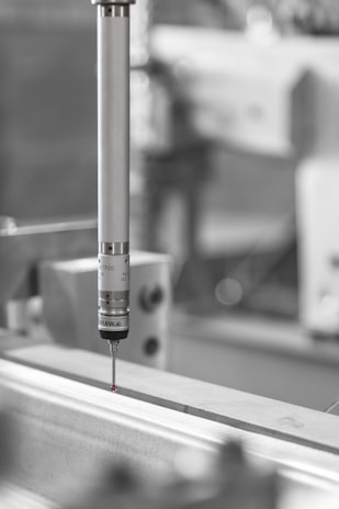 A precision measuring probe is positioned above a metallic surface, likely part of an industrial machine or CNC equipment. The focus is primarily on the metallic probe with a small red tip, highlighting its use in detailed measurements. The background is blurred, emphasizing an industrial setting.