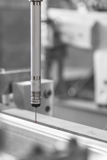 A precision measuring probe is positioned above a metallic surface, likely part of an industrial machine or CNC equipment. The focus is primarily on the metallic probe with a small red tip, highlighting its use in detailed measurements. The background is blurred, emphasizing an industrial setting.