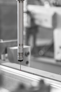 A precision measuring probe is positioned above a metallic surface, likely part of an industrial machine or CNC equipment. The focus is primarily on the metallic probe with a small red tip, highlighting its use in detailed measurements. The background is blurred, emphasizing an industrial setting.