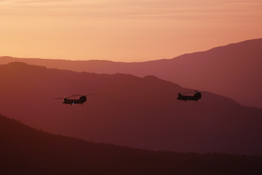 two military helicopters flying over a mountain range