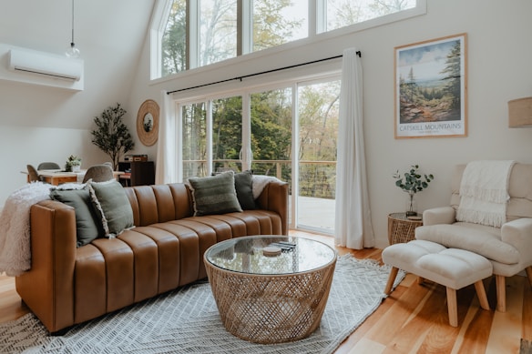 A cozy and modern living room features a brown leather sofa with green cushions, a round woven coffee table, and a geometric-patterned rug. A soft beige armchair with a footrest is placed near a large window that overlooks a forested area. The room is decorated with a Catskill Mountains poster, indoor plants, and a dining table in the background. Ample natural light fills the space, creating a warm and inviting atmosphere.
