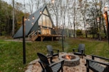 A modern A-frame house with large windows is set in a forested area. In the foreground, a circular stone fire pit is surrounded by black Adirondack chairs. String lights hang above the scene, adding a warm glow to the tranquil forest setting.