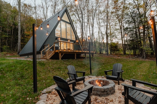 A modern A-frame house with large windows is set in a forested area. In the foreground, a circular stone fire pit is surrounded by black Adirondack chairs. String lights hang above the scene, adding a warm glow to the tranquil forest setting.