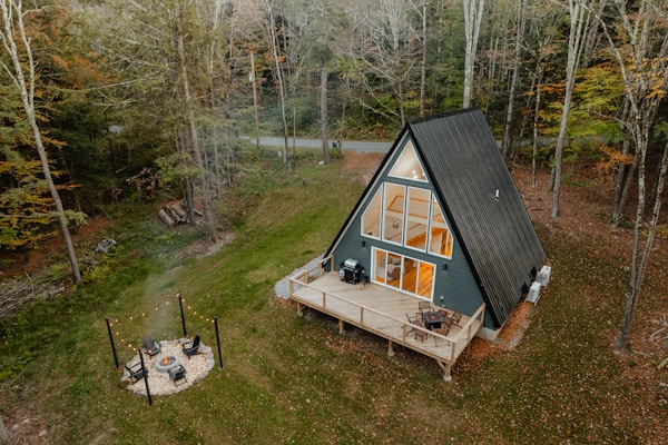 A secluded A-frame cabin with large triangular windows stands surrounded by a dense forest during autumn. The cabin features a wooden deck with outdoor furniture and a barbecue grill. In the foreground, a small fire pit is encircled by chairs and string lights, creating a cozy outdoor setting.