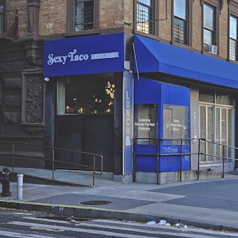A corner restaurant with a blue awning and signage reading 'Sexy Taco Dirty Cash'. The building is situated at a street corner with a sidewalk and ramp. Large windows display interior lighting, and smaller text on the windows mentions catering and delivery options. The exterior has a mix of brick and stonework.