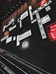A collection of various illuminated signs and logos on a wall in a dimly lit environment, including phrases and drawings in different fonts and styles. An escalator is visible in the foreground, contributing to an urban, indoor setting.