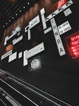 A collection of various illuminated signs and logos on a wall in a dimly lit environment, including phrases and drawings in different fonts and styles. An escalator is visible in the foreground, contributing to an urban, indoor setting.