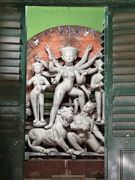 A detailed clay sculpture of a multi-armed goddess figure surrounded by smaller figures, depicting a mythological scene. The central figure stands with impressive poise, holding various items in her hands. The sculpture includes a fierce lion and another figure at the base, emerging from a green backdrop with wooden shutter panels framing the scene.