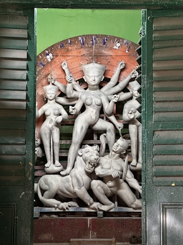 A detailed clay sculpture of a multi-armed goddess figure surrounded by smaller figures, depicting a mythological scene. The central figure stands with impressive poise, holding various items in her hands. The sculpture includes a fierce lion and another figure at the base, emerging from a green backdrop with wooden shutter panels framing the scene.