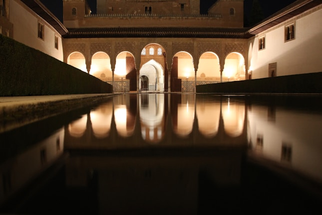 An illuminated courtyard featuring an architectural structure with elegant arches and intricate details, reflecting in a long, tranquil pool of water. It's night-time, and the soft warm lights create a serene atmosphere.
