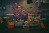 An outdoor flower stall is set up against the backdrop of a colorful mural featuring a peacock feather design with vibrant purples, blues, and yellows. Multiple garlands of flowers hang from a rack, and the area is illuminated with warm lighting.
