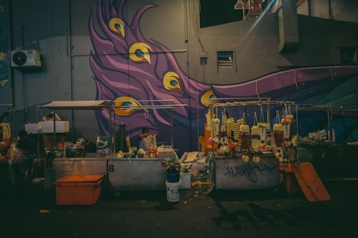 An outdoor flower stall is set up against the backdrop of a colorful mural featuring a peacock feather design with vibrant purples, blues, and yellows. Multiple garlands of flowers hang from a rack, and the area is illuminated with warm lighting.