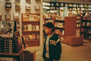 A young person in a jacket stands inside a bookstore, looking at a display featuring Marvel comics. The store has numerous shelves filled with books, creating a cozy and intellectual atmosphere.